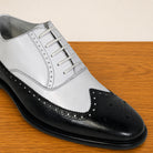 A men's wingtip oxford shoe in a two-tone black and white leather finish, displayed on a wooden surface against a light beige background.