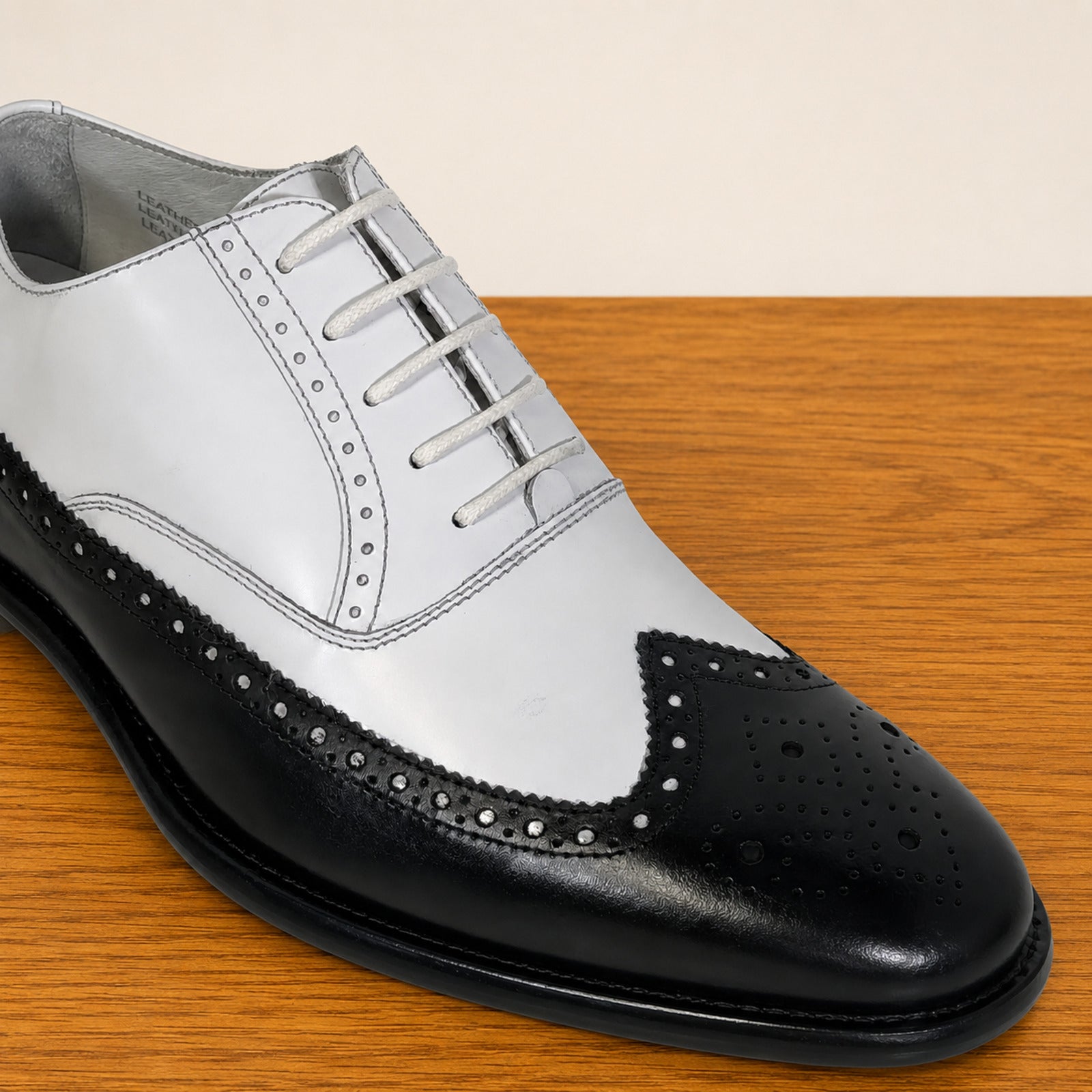 A men's wingtip oxford shoe in a two-tone black and white leather finish, displayed on a wooden surface against a light beige background.