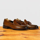 A pair of Dark Caramel leather tassel loafers with dark brown stacked heels, displayed on a wooden surface against a light gray background.