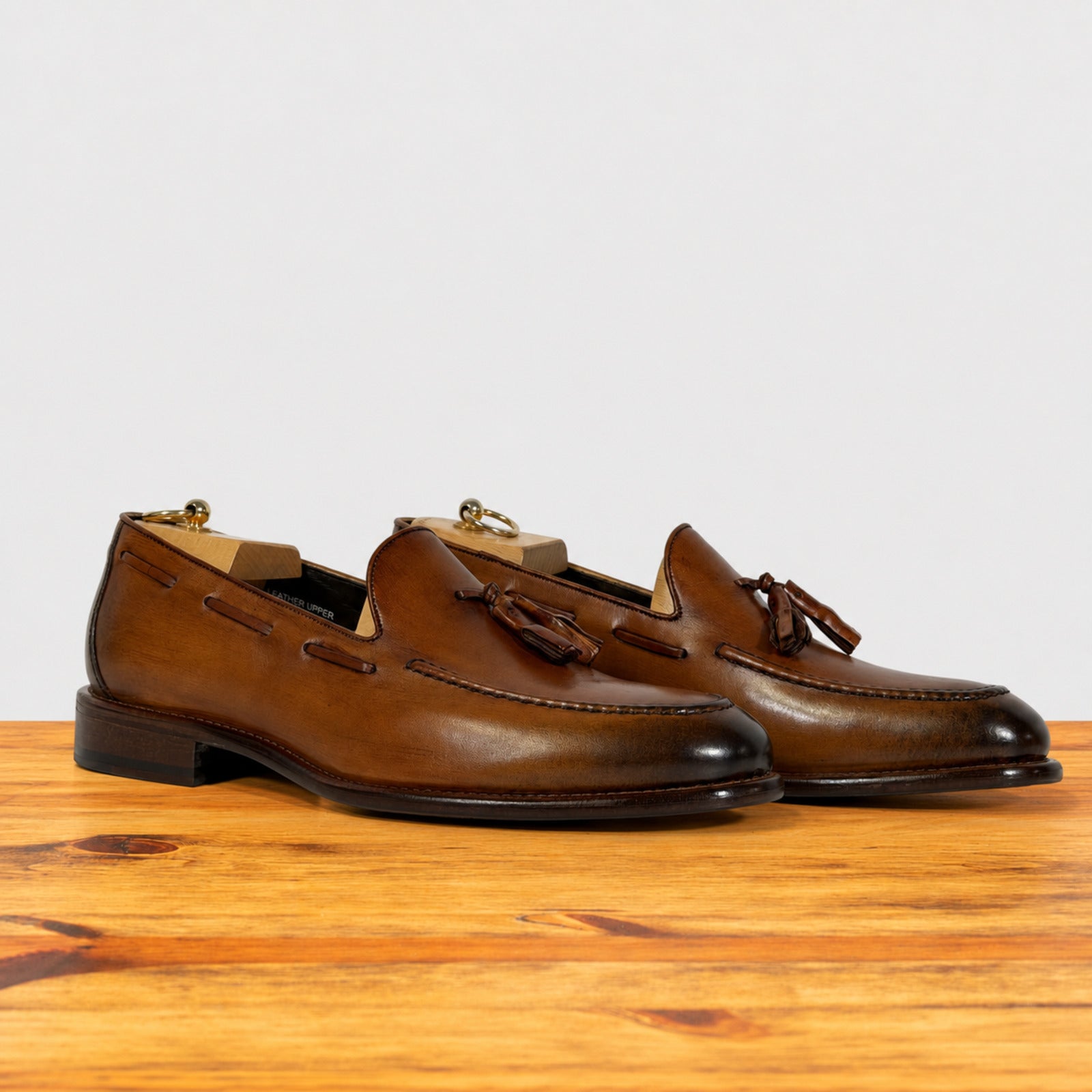 A pair of Dark Caramel leather tassel loafers with dark brown stacked heels, displayed on a wooden surface against a light gray background.