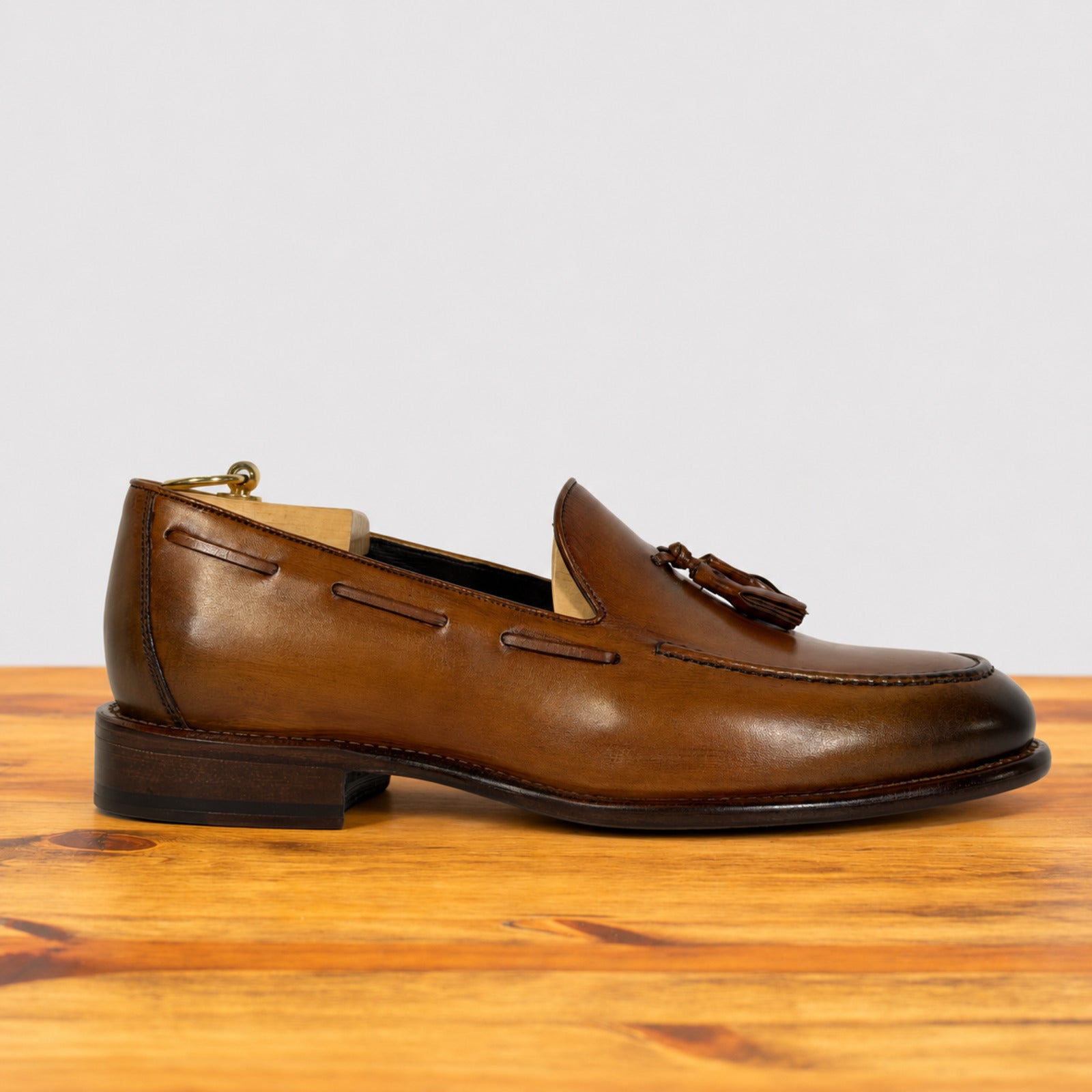 A single Dark Caramel  leather tassel loafer shoe resting on a wooden surface against a light gray background.