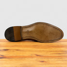 The underside of a brown leather shoe sole, showing the heel block and toe cap, resting on a wooden surface against a light gray background.