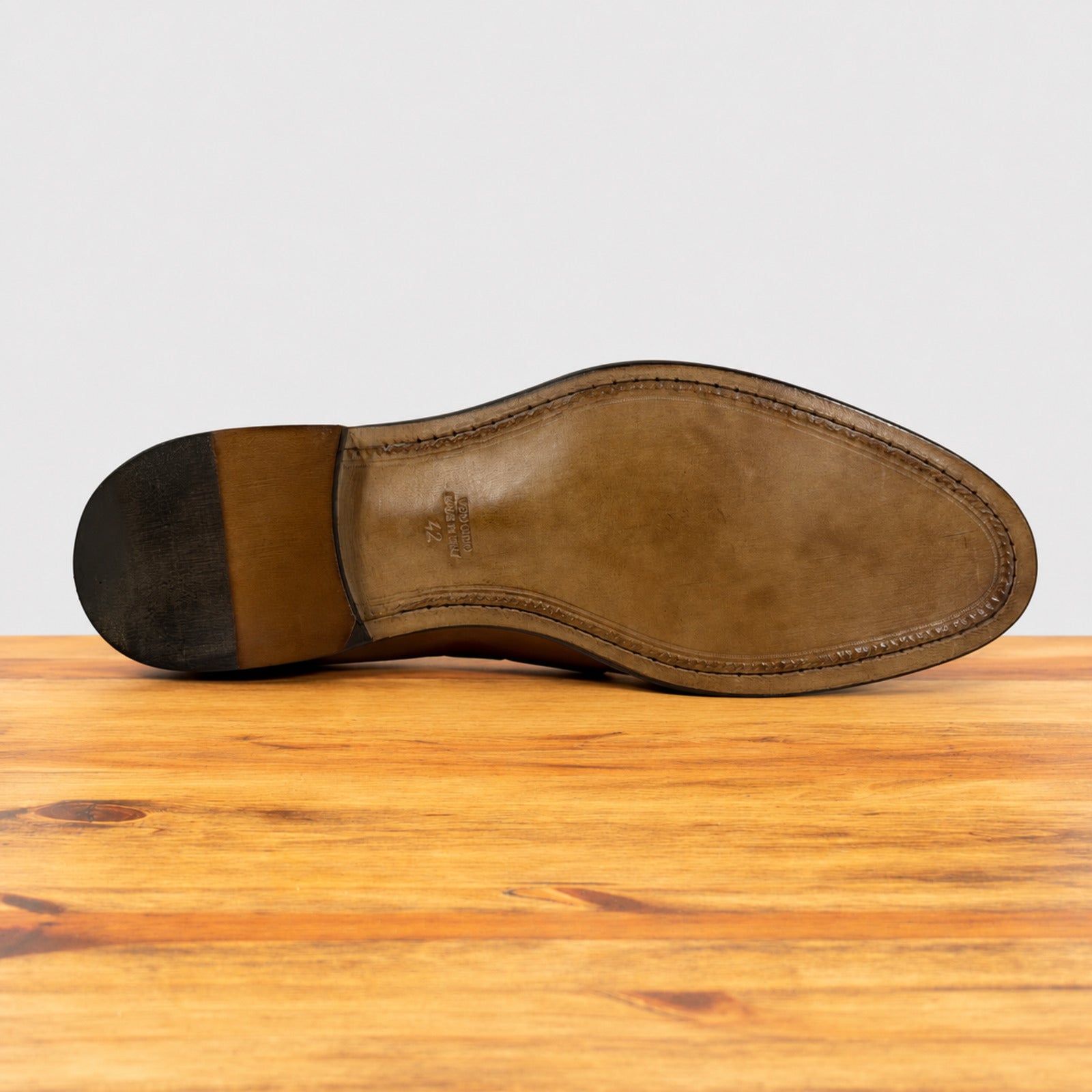 The underside of a brown leather shoe sole, showing the heel block and toe cap, resting on a wooden surface against a light gray background.