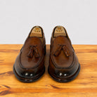 A pair of Dark Caramel leather tassel loafers with polished toes, displayed on wooden shoe trees on a wooden surface against a light gray background.