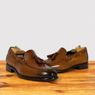 A pair of men's Dark Caramel leather tassel loafers with dark brown pointed toes and low stacked heels, displayed on a wooden surface against a light gray background.