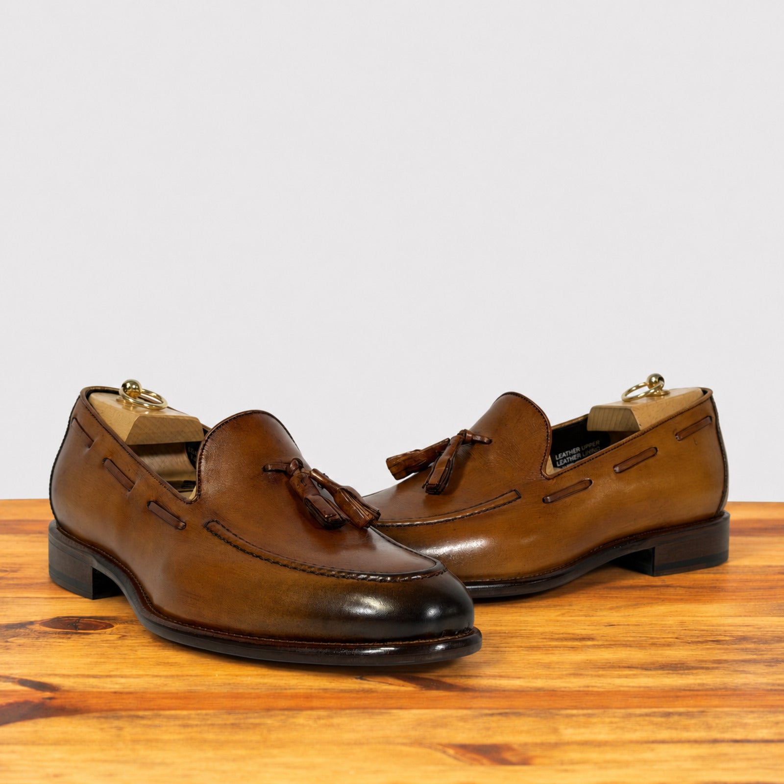 A pair of men's Dark Caramel leather tassel loafers with dark brown pointed toes and low stacked heels, displayed on a wooden surface against a light gray background.
