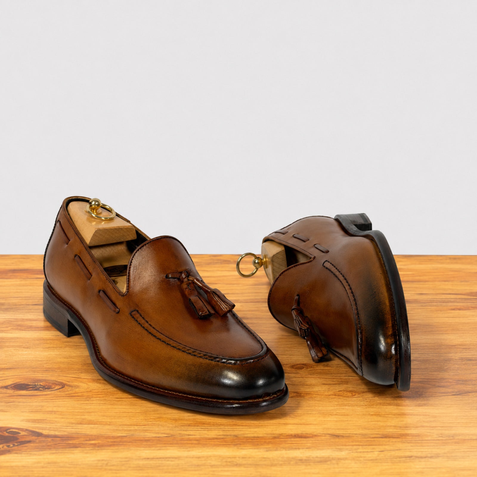 A pair of Dark Caramel leather tassel loafers displayed on a wooden surface against a light gray background. One shoe is shown from the front, revealing the tassel and side profile, while the other is turned to show the inner side and heel.