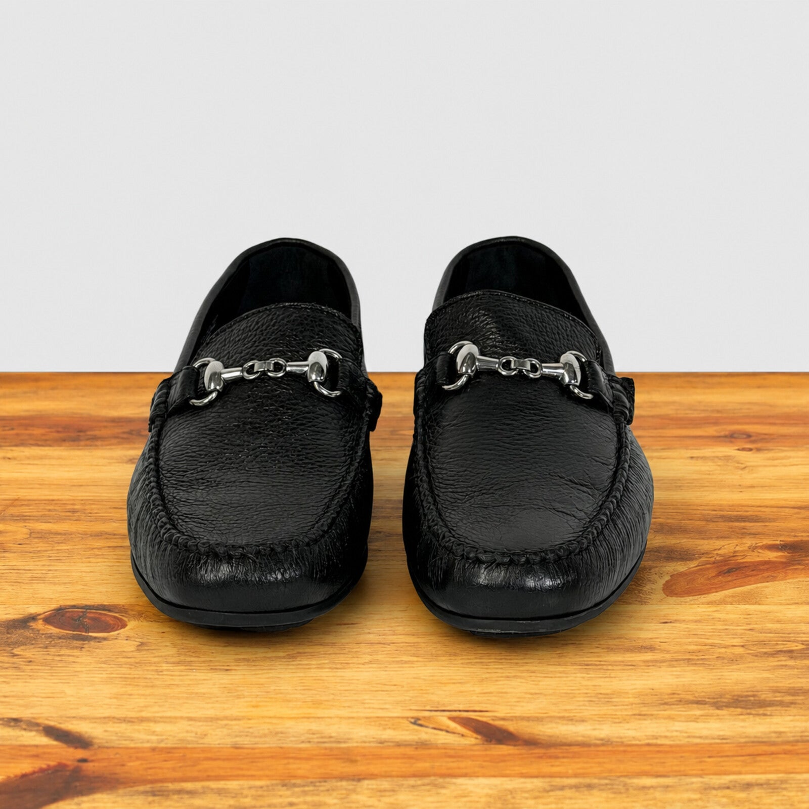 A pair of black leather moccasin-style loafers with silver strap, displayed on a wooden surface against a light gray background.