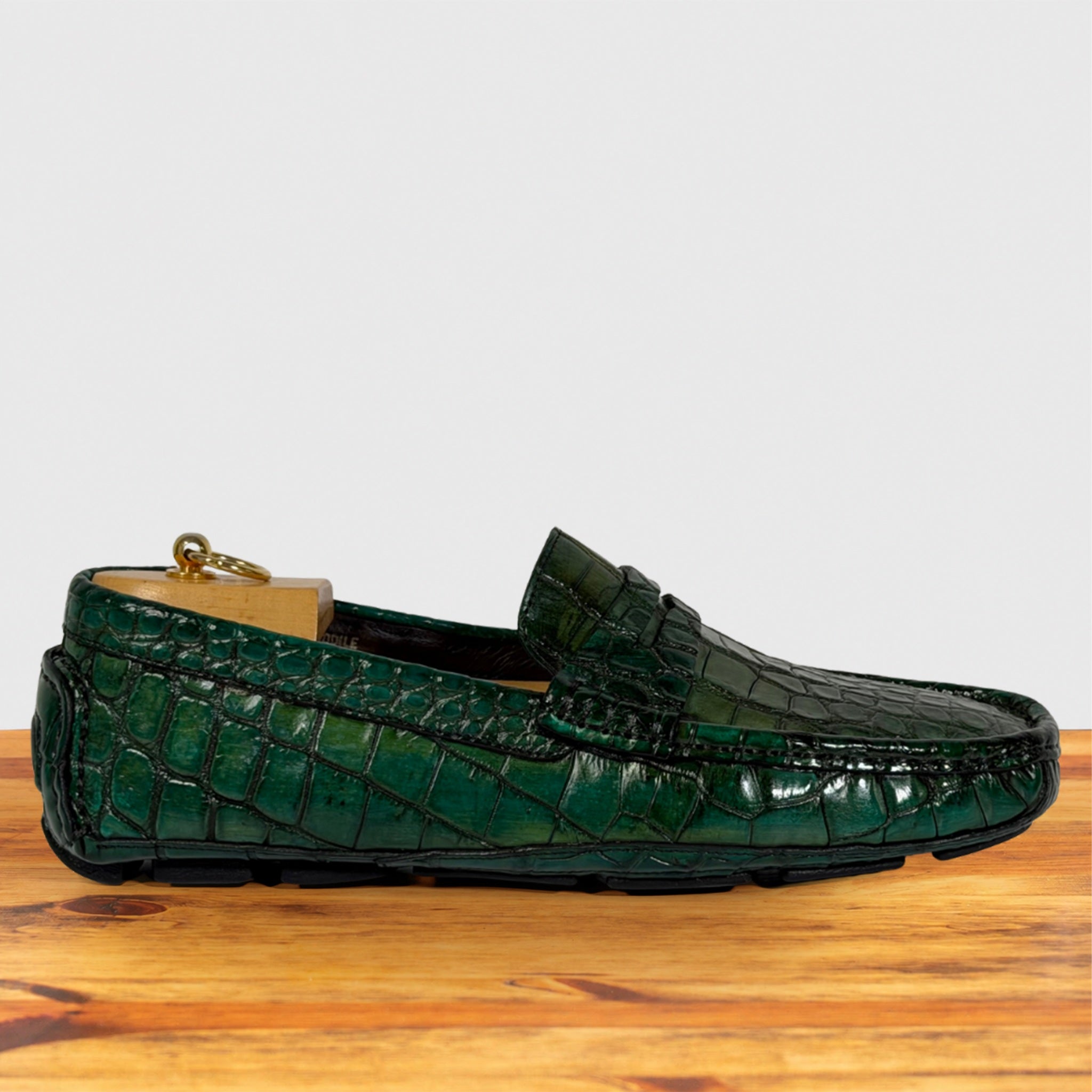 A single green Genuine Crocodile loafer driving shoe resting on a wooden surface against a light gray background.