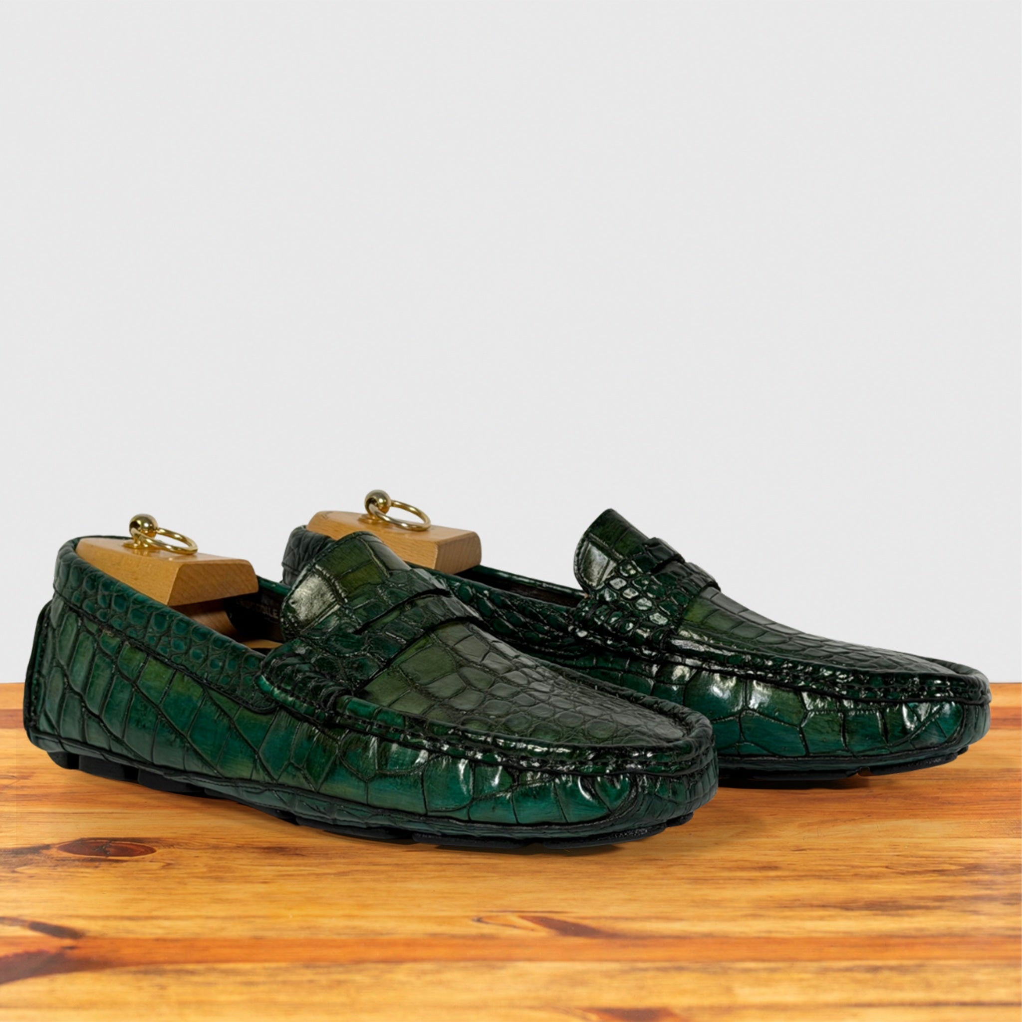 A pair of Emerald Green Genuine Crocodile leather penny loafers resting on a wooden surface with wooden shoe trees inside, against a plain light gray background.