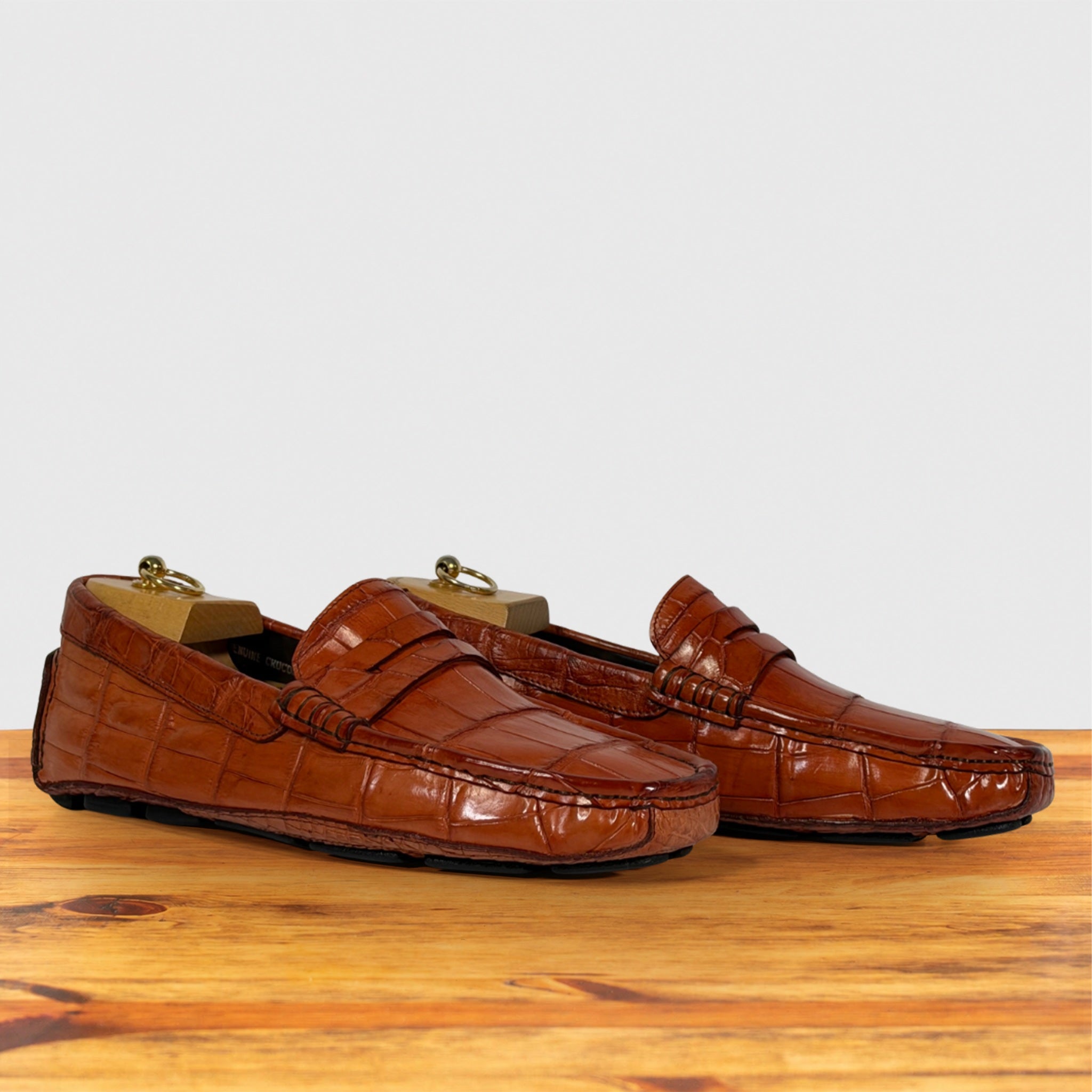 A pair of Burnt Orange Genuine Crocodile leather penny loafers, displayed on a wooden surface against a light gray background.