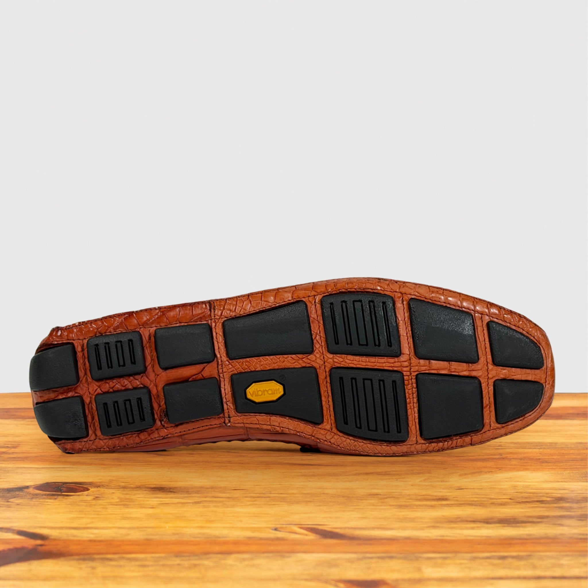 The outsole of a brown Genuine Crocodile leather shoe resting on a wooden surface against a light gray background.