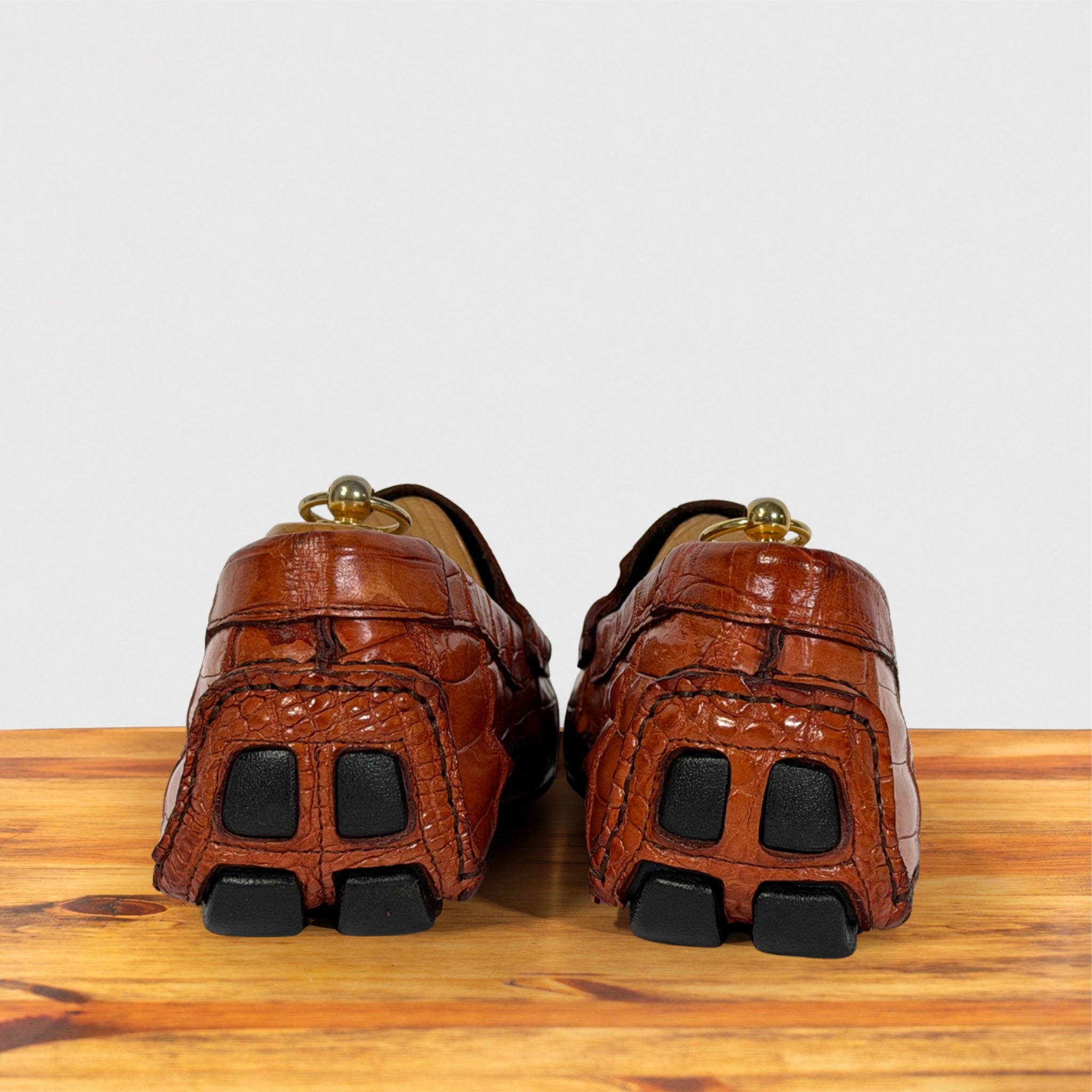 A pair of Burnt Orange Genuine Crocodile leather loafers shown from the back, resting on a wooden surface against a light gray background.