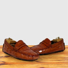 A pair of Burnt Orange crocodile-embossed leather penny loafers resting on a wooden surface against a light gray background.