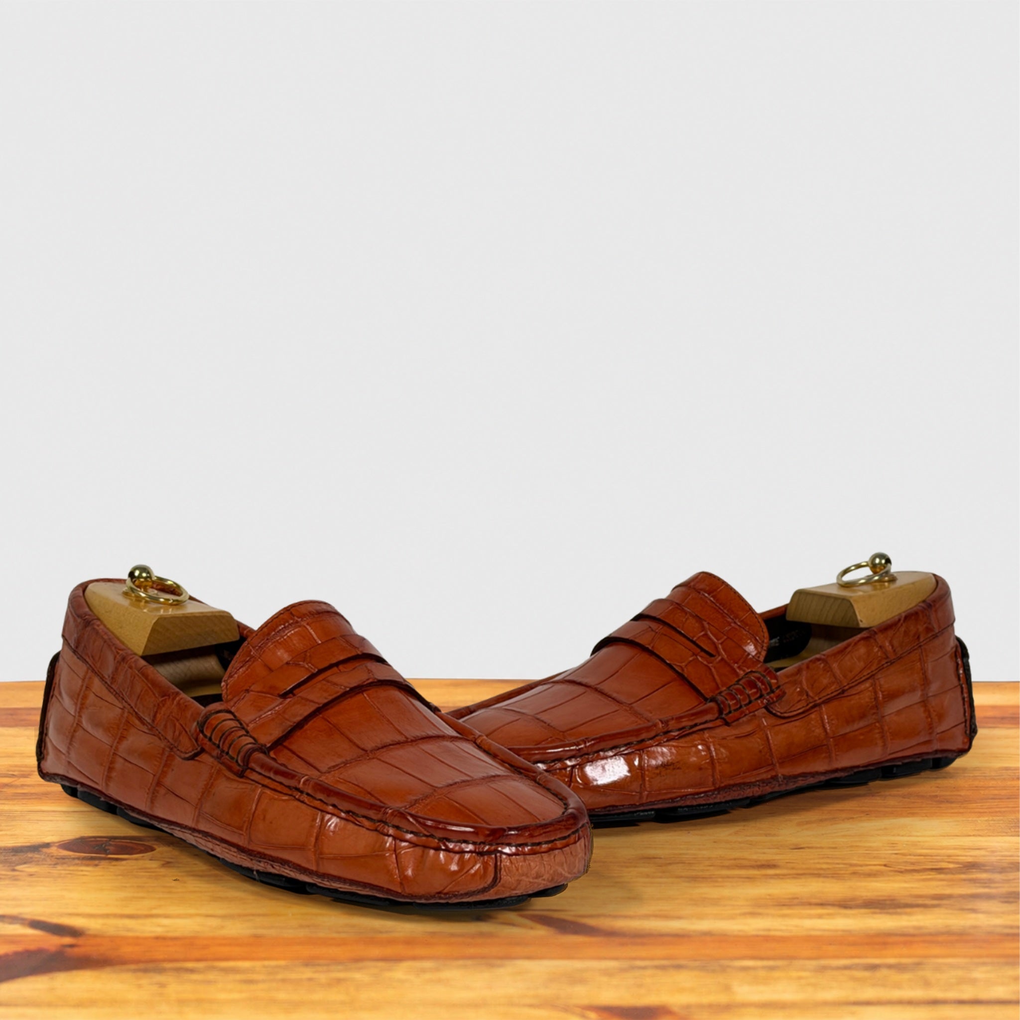 A pair of Burnt Orange crocodile-embossed leather penny loafers resting on a wooden surface against a light gray background.