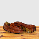 A pair of Burnt Orange Genuine Crocodile leather penny loafers displayed on a wooden surface against a light gray background.