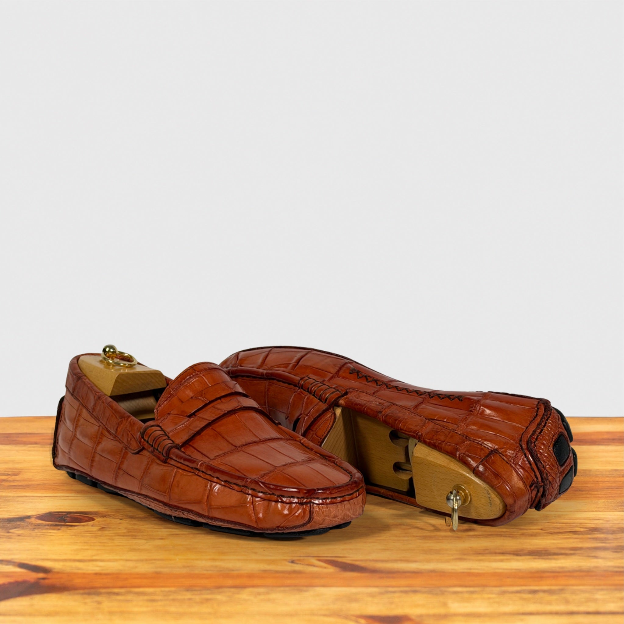 A pair of Burnt Orange Genuine Crocodile leather penny loafers displayed on a wooden surface against a light gray background.