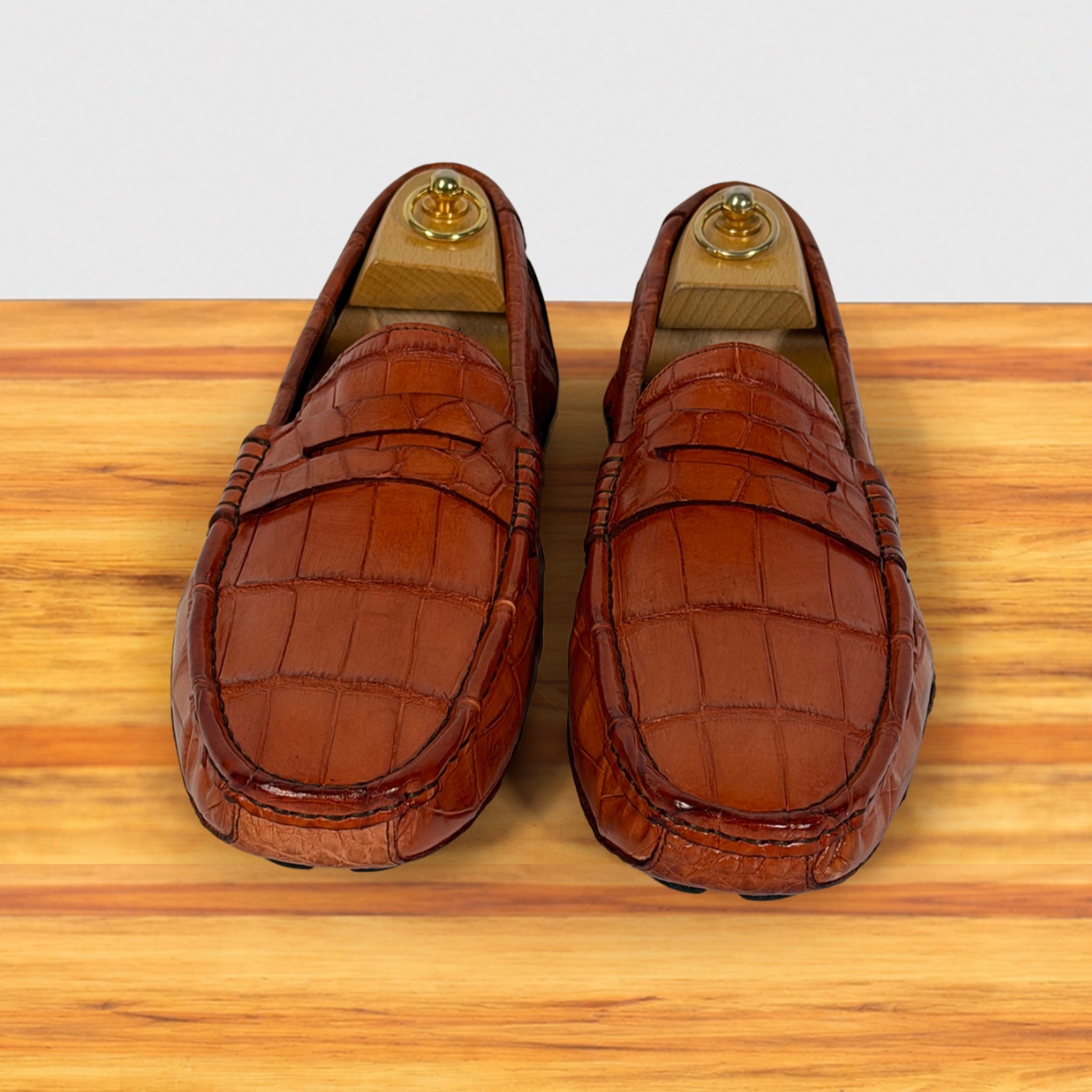 A pair of Burnt Orange Genuine Crocodile leather penny loafers resting on a light wood surface against a white background.