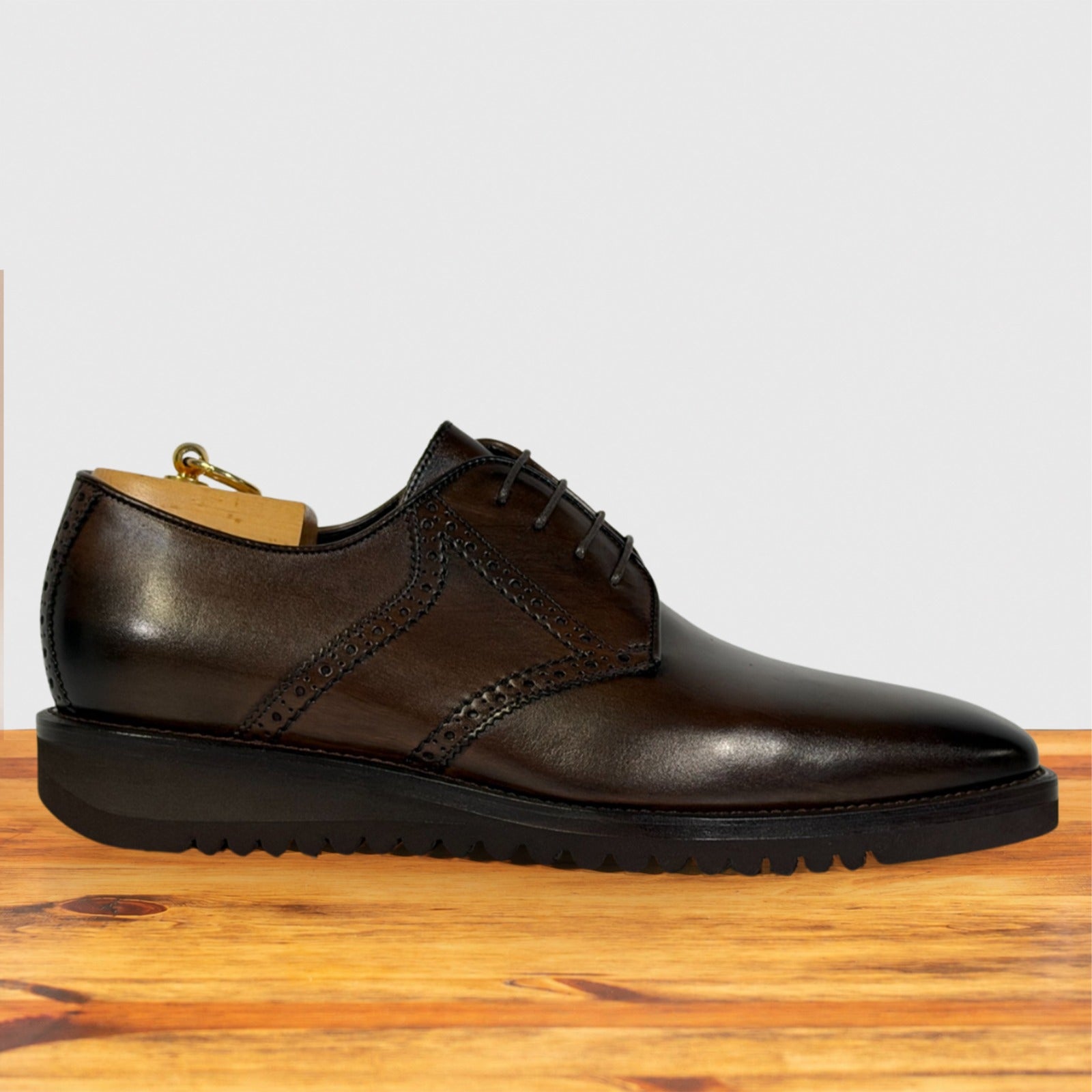 A moor leather lace-up dress shoe with brogue detailing and a thick black sole, displayed on a wooden surface against a light gray background.
