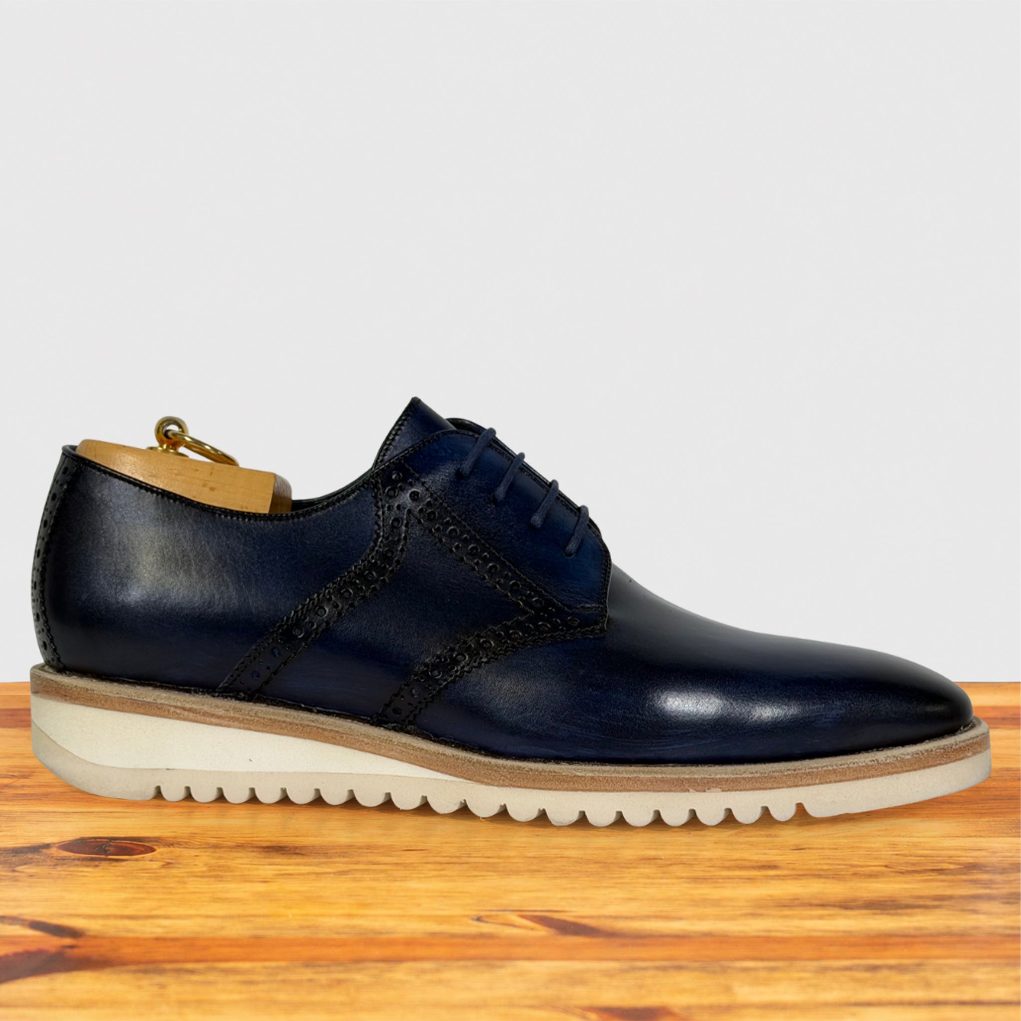A navy blue leather lace-up dress shoe with a cream rubber sole, displayed on a wooden surface against a light gray background.