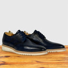 A pair of navy blue leather oxford shoes with brogue detailing and white lug soles, displayed on a wooden surface against a light gray background.