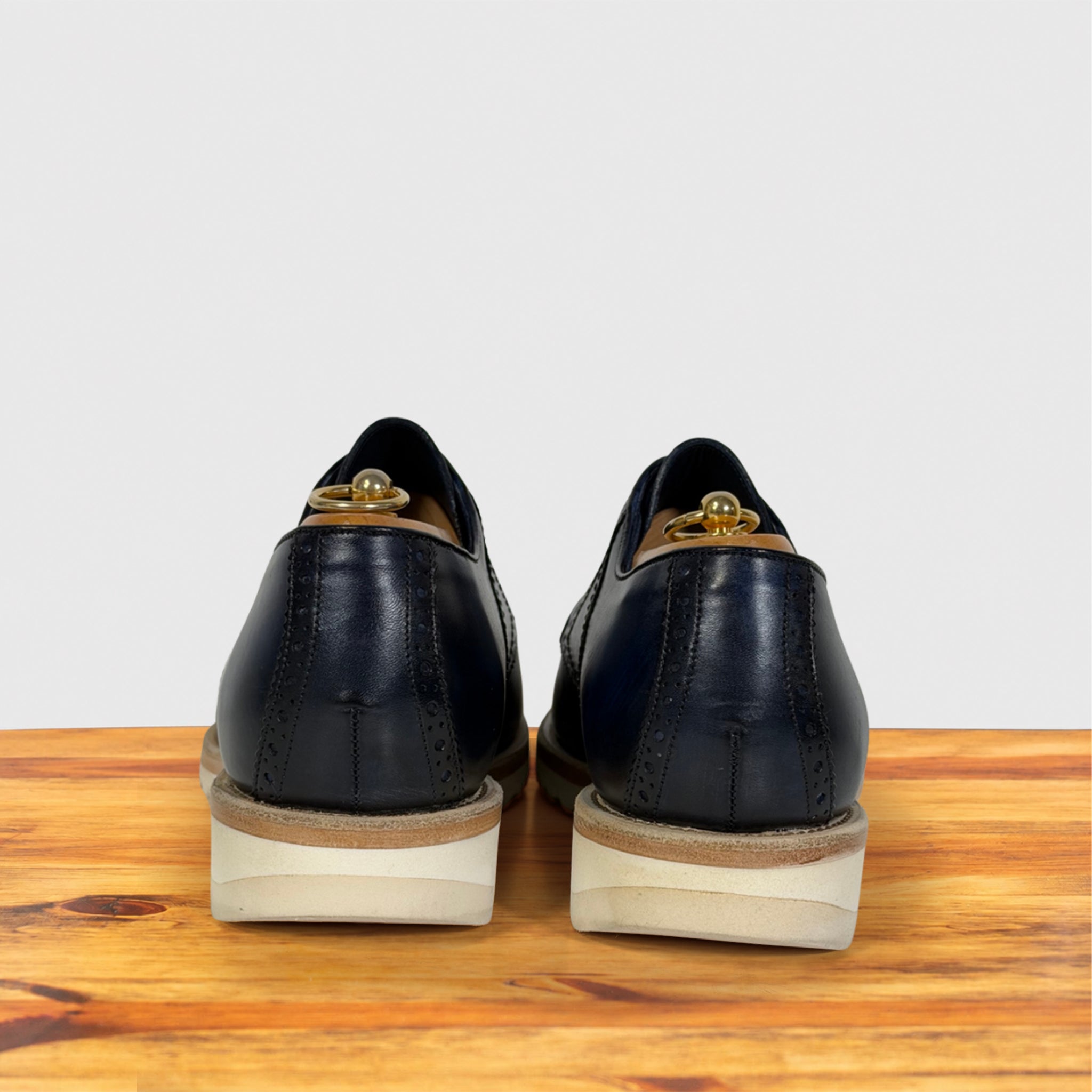 A pair of dark navy blue leather wingtip oxford shoes shown from the back, resting on a wooden surface against a light gray background.