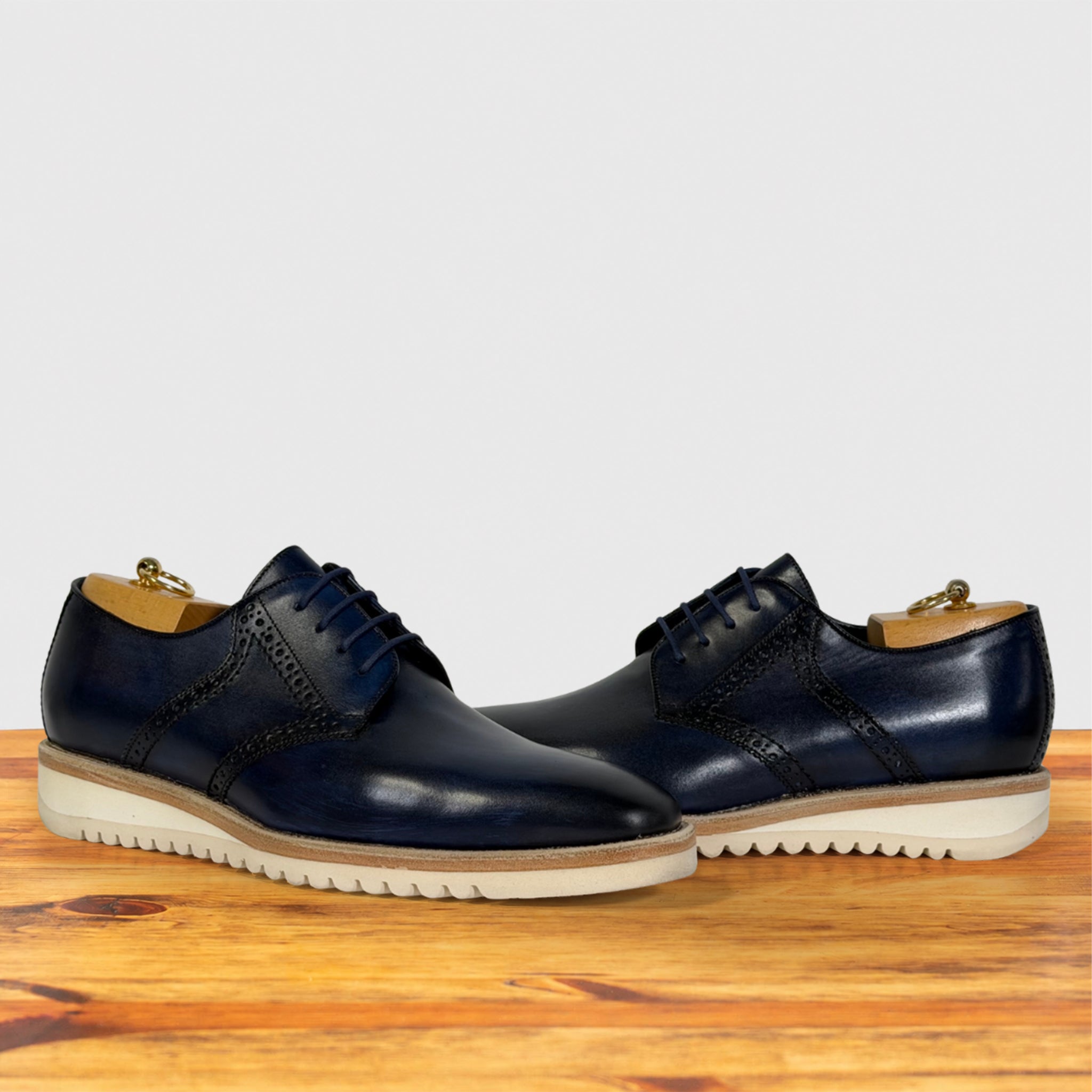A pair of navy blue leather lace-up dress shoes with brogue detailing and white wavy soles, displayed on a wooden surface against a light gray background.