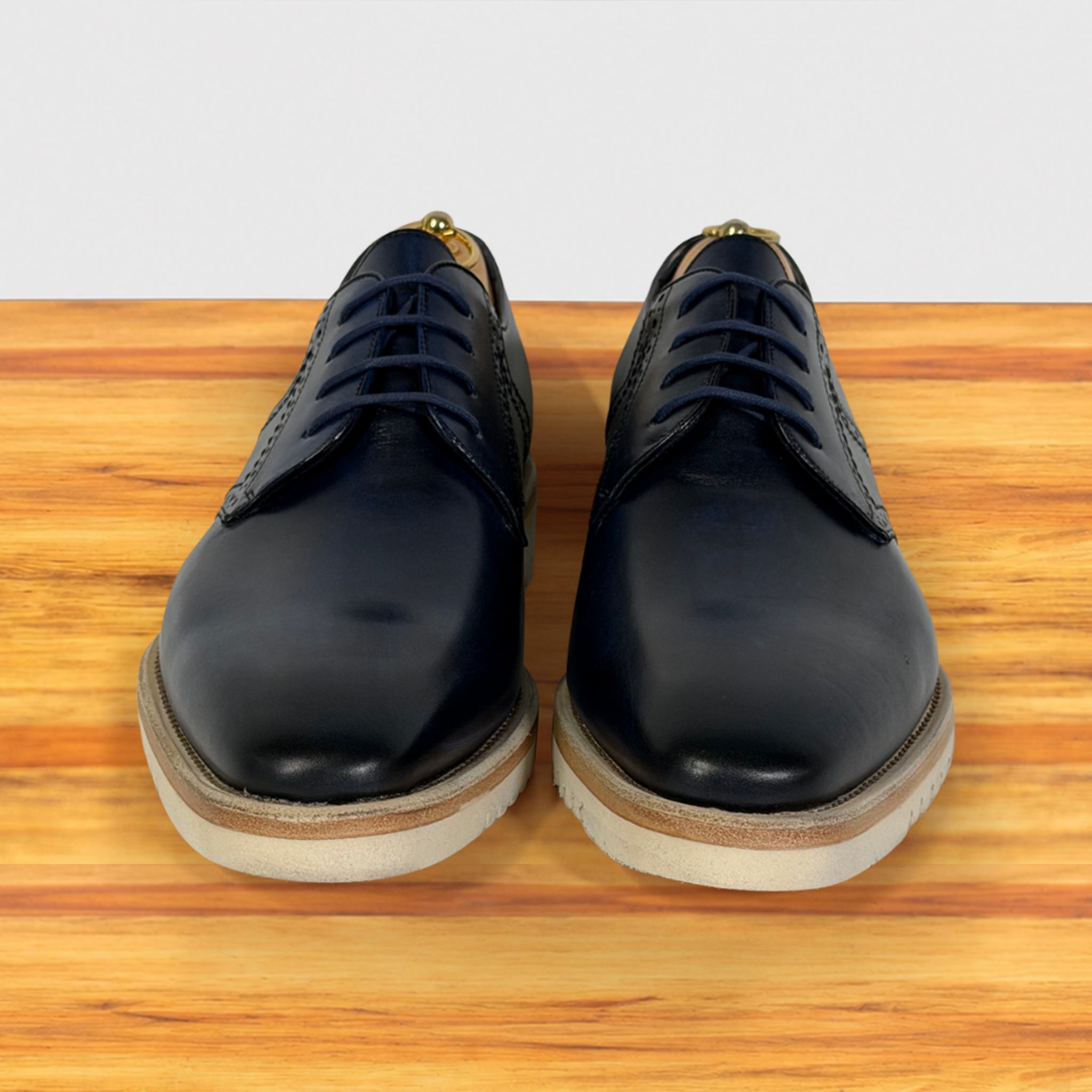A pair of men's blue leather oxford shoes with brogue detailing, shown from the front on a wooden surface against a light gray background.