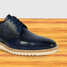 A dark navy blue leather lace-up dress shoe with decorative brogue perforations and a thick cream-colored sole, shown on a wooden surface against a light gray background.