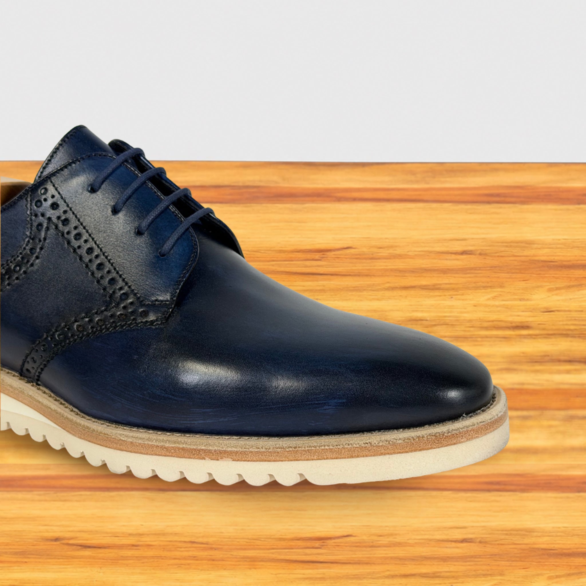 A dark navy blue leather lace-up dress shoe with decorative brogue perforations and a thick cream-colored sole, shown on a wooden surface against a light gray background.