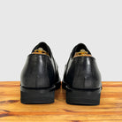 A pair of black leather dress shoes shown from the back, resting on a wooden surface against a light gray background.