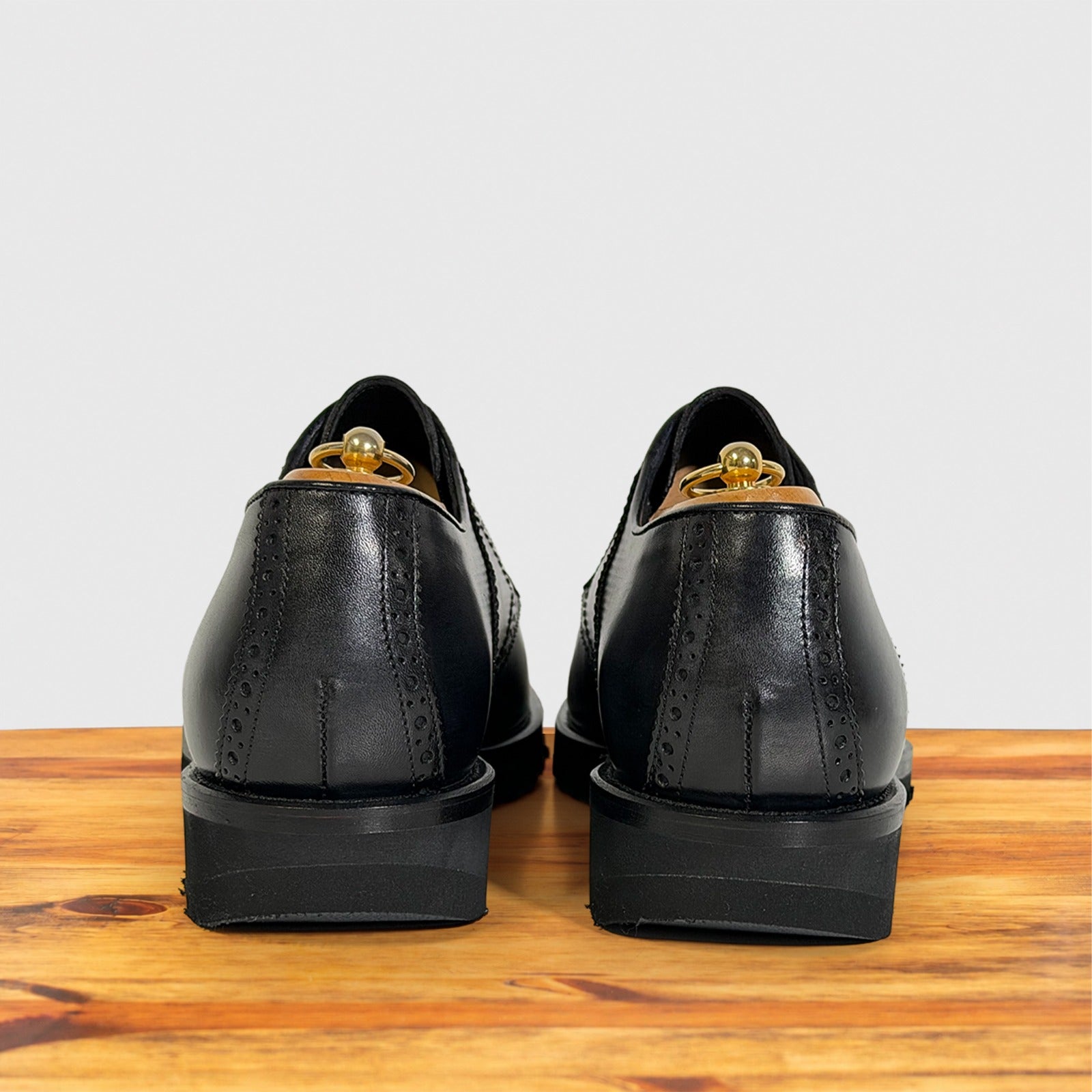 A pair of black leather dress shoes shown from the back, resting on a wooden surface against a light gray background.