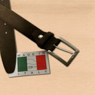 A dark brown suede belt with a silver metal buckle, displayed on a light wood background next to a silver 'MADE IN ITALY' tag featuring the Italian flag.