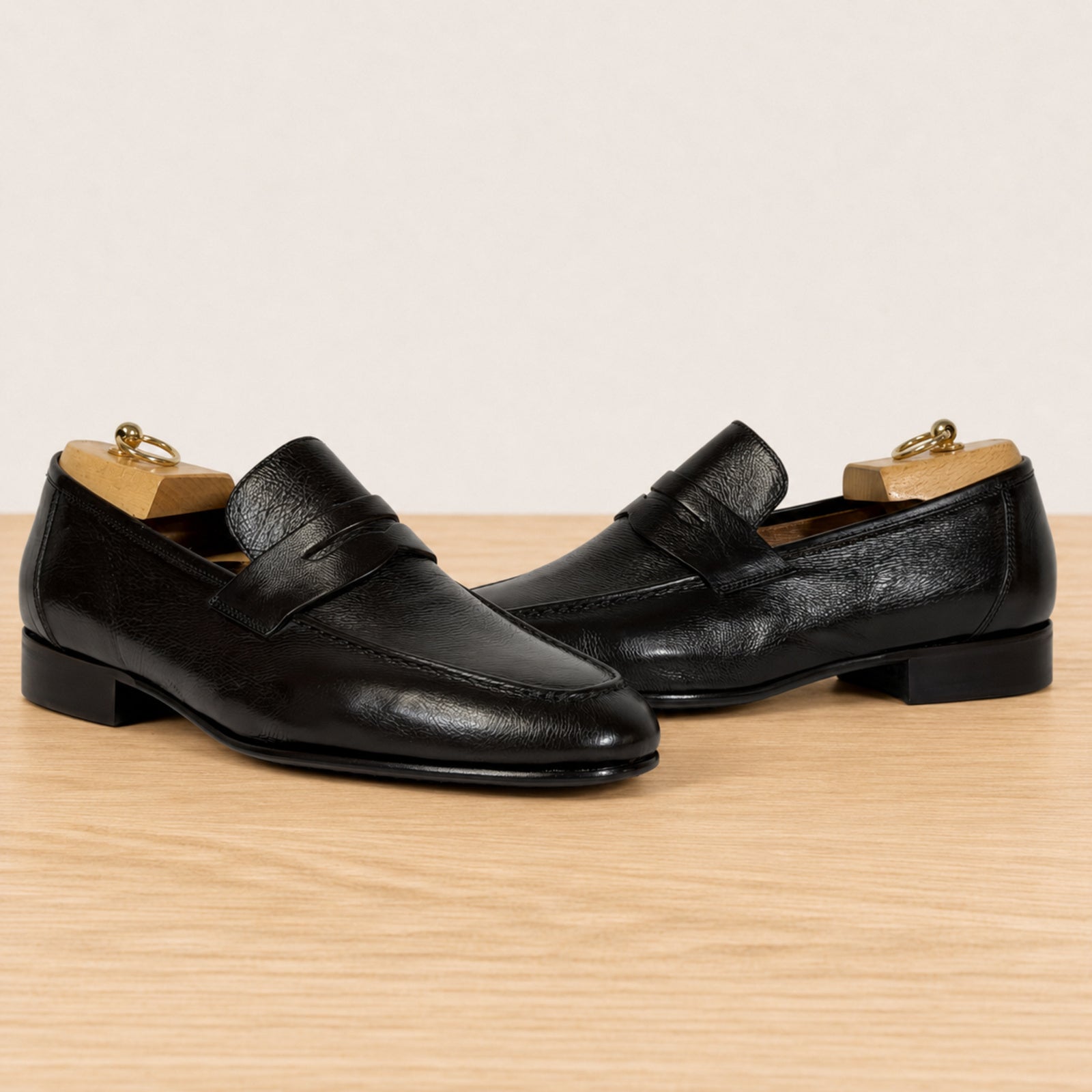 A pair of dark brown leather penny loafers with a subtle textured grain, displayed on a light wood surface against a warm off-white background.