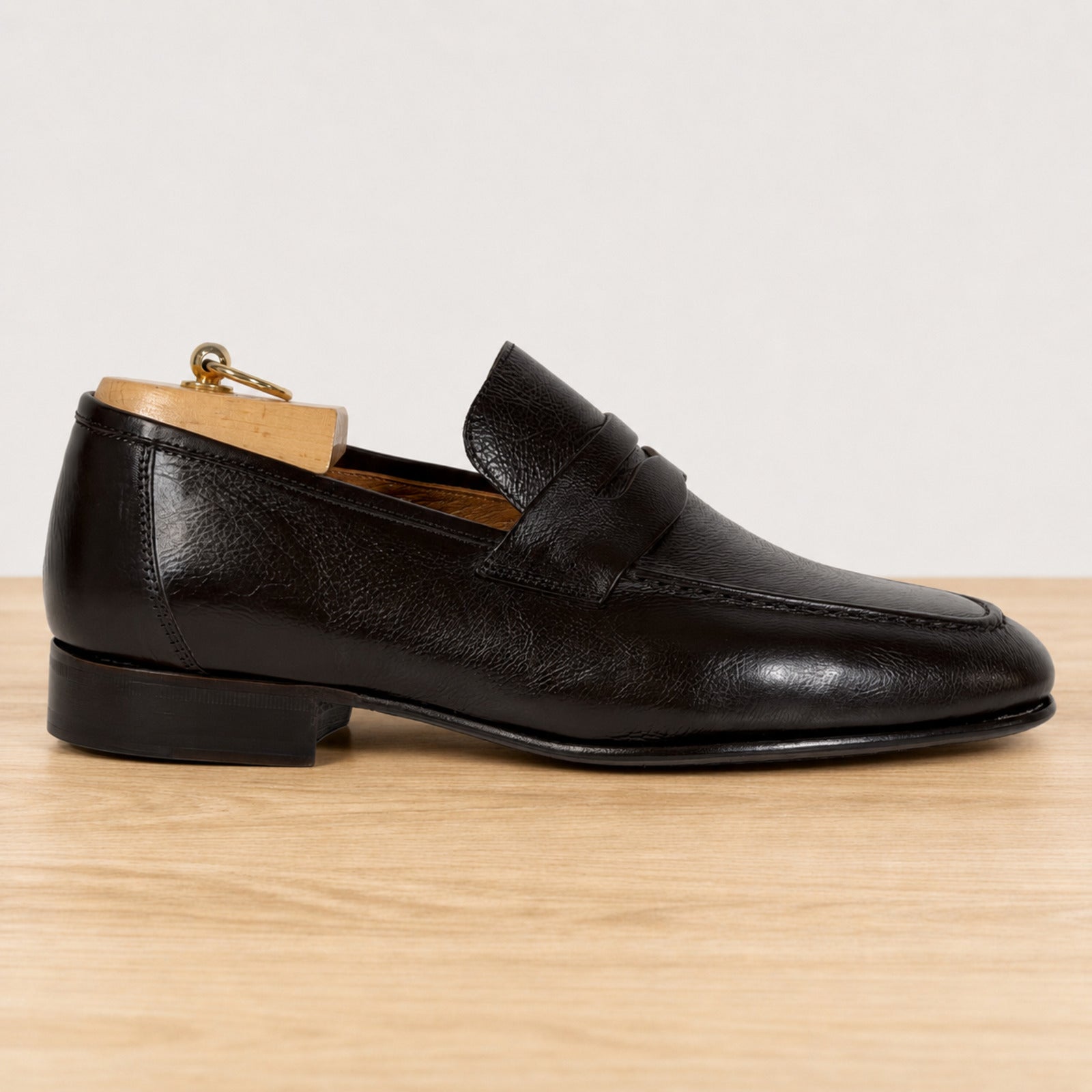 A single dark brown leather penny loafer with a pebbled texture, shown in profile on a light wood surface against a white background. A wooden shoe tree is inserted inside the shoe.