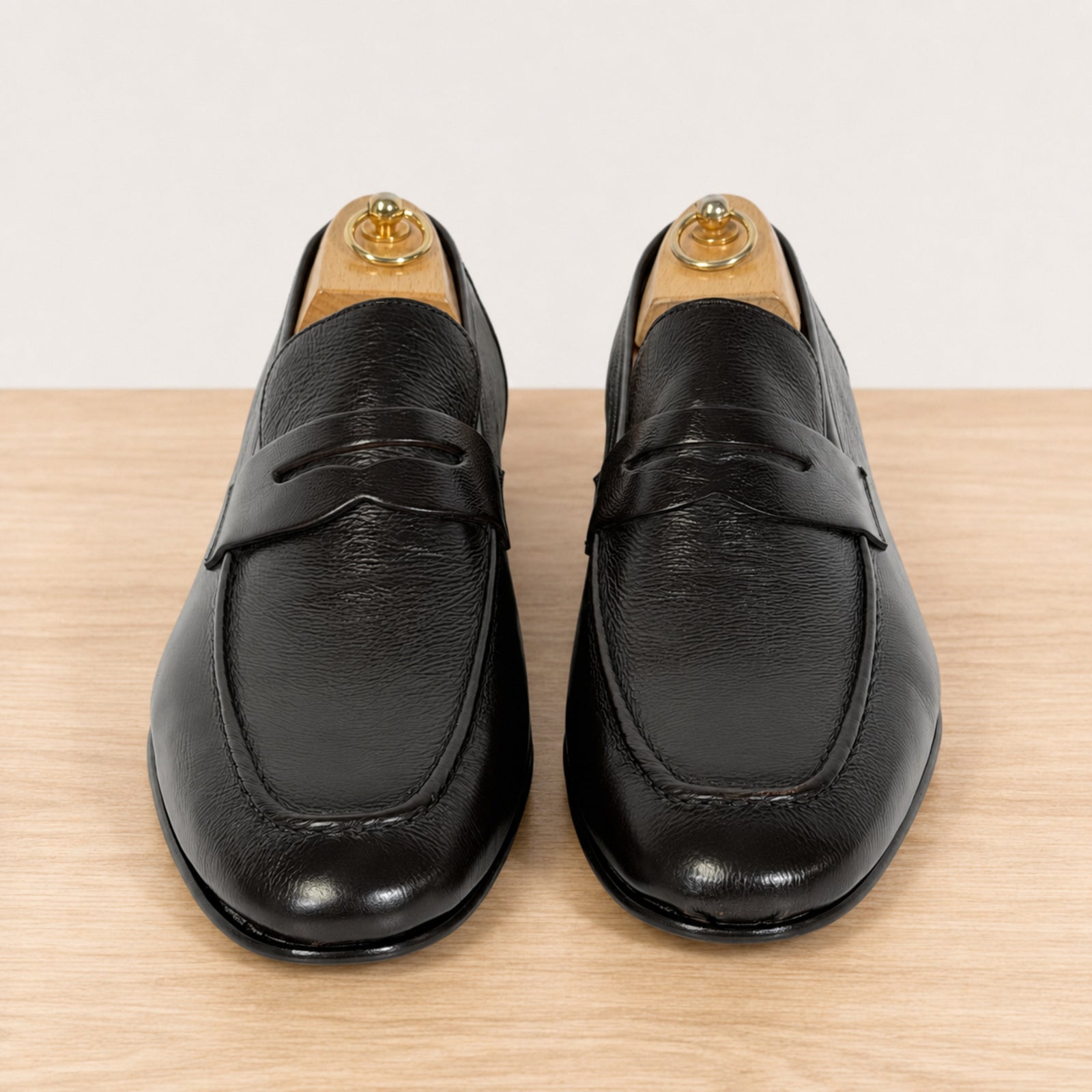 A pair of dark brown leather penny loafers with a pebbled texture, shown on wooden shoe trees against a light wood and white background.
