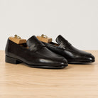 A pair of dark brown leather penny loafers with wooden shoe trees inserted, displayed on a light wood surface against a plain light gray background.