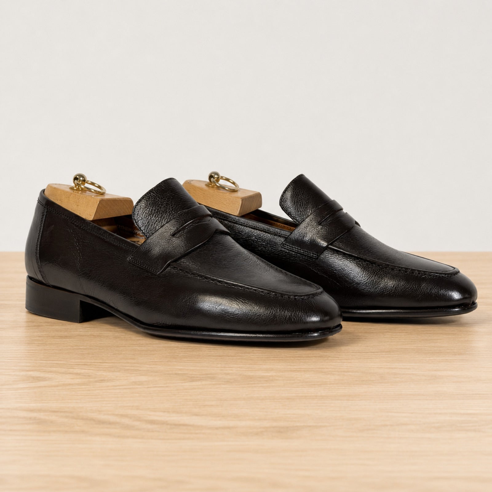 A pair of dark brown leather penny loafers with wooden shoe trees inserted, displayed on a light wood surface against a plain light gray background.