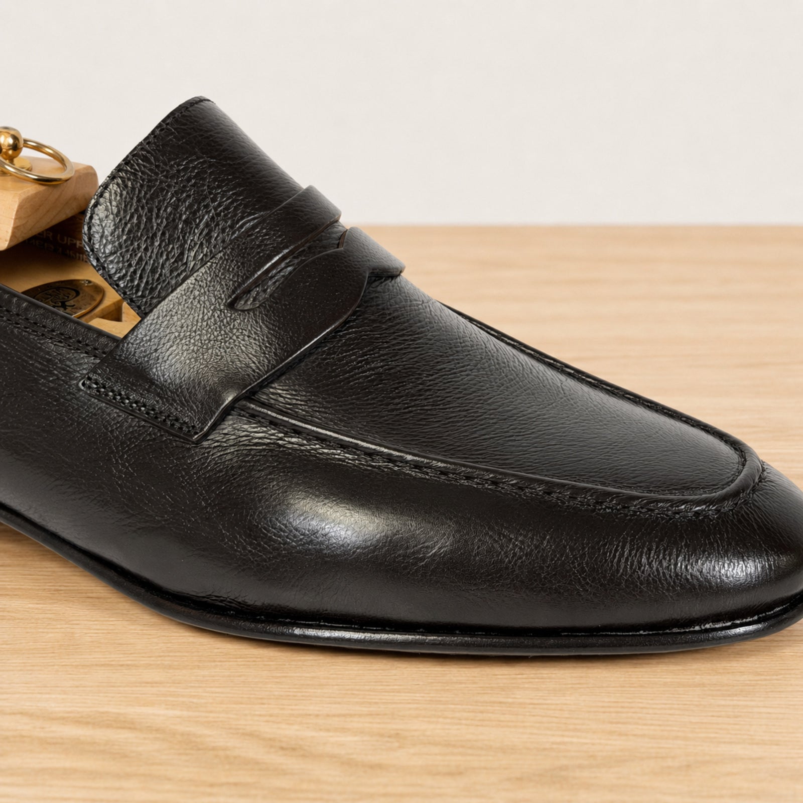 A close-up of a dark brown leather penny loafer on a wooden shoe tree, set against a light wood tabletop and a neutral background.