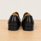 A pair of dark brown leather loafers shown from the back, resting on a light wood surface against a plain off-white background.