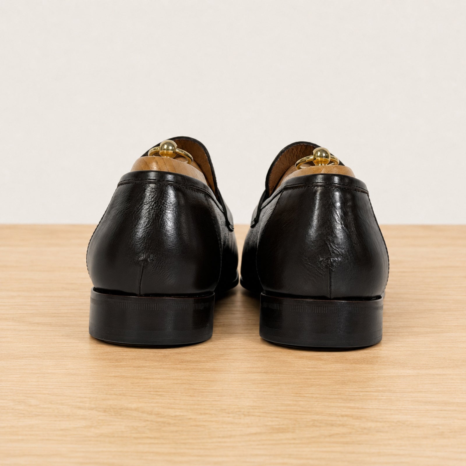 A pair of dark brown leather loafers shown from the back, resting on a light wood surface against a plain off-white background.