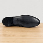 The outsole view of a black leather dress shoe, showing the toe, heel, and stitching details, resting on a light wood surface against a neutral gray background.