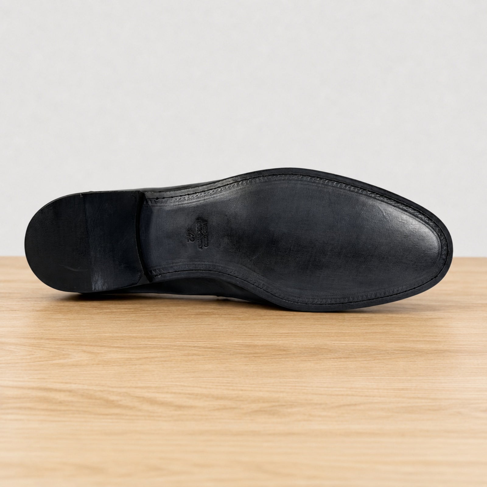 The outsole view of a black leather dress shoe, showing the toe, heel, and stitching details, resting on a light wood surface against a neutral gray background.