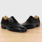 A pair of black leather penny loafers with low stacked heels, displayed on wooden shoe trees on a light wood surface against a white background.