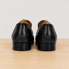 A pair of black leather loafers shown from the back, resting on a light wood surface against a plain light gray background.