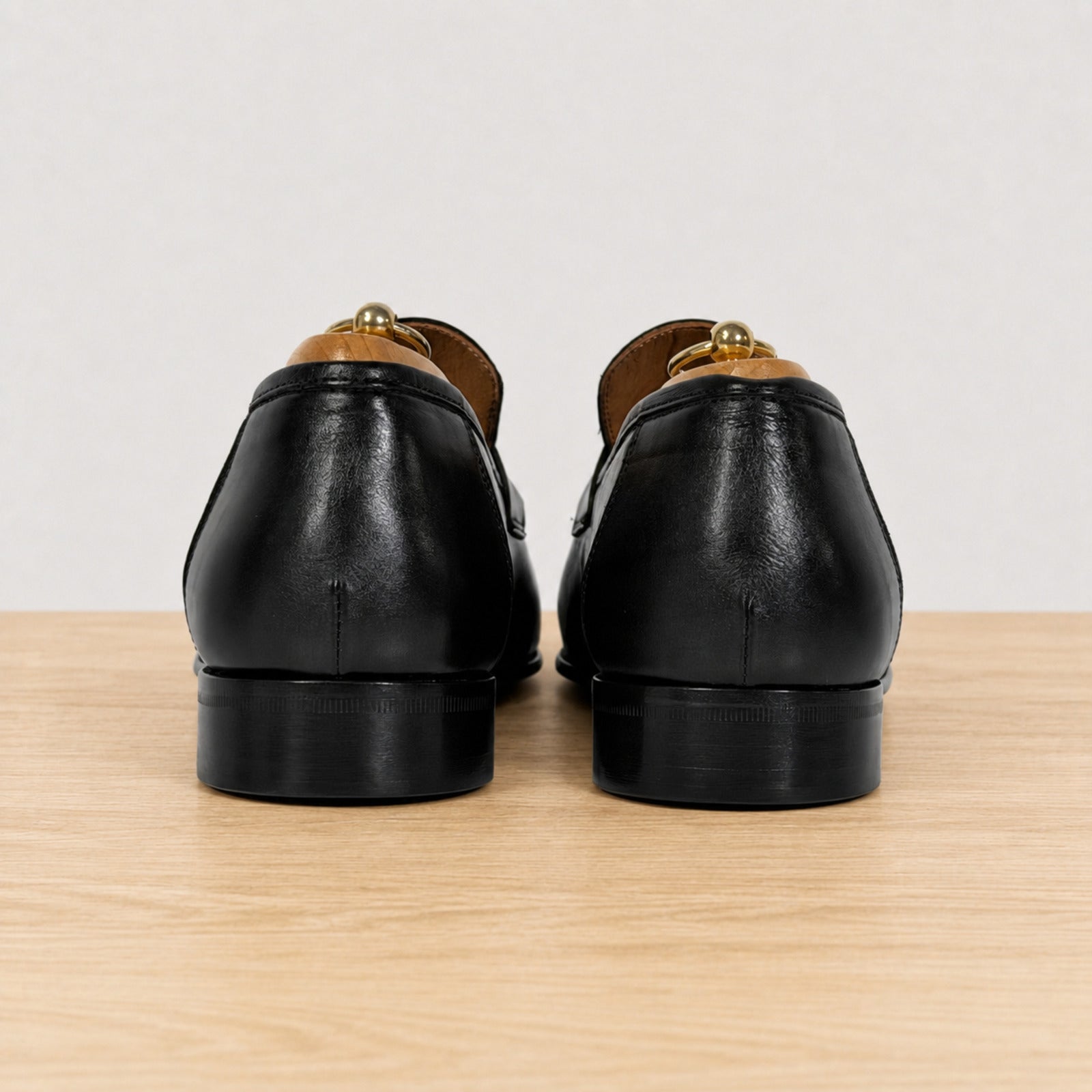 A pair of black leather loafers shown from the back, resting on a light wood surface against a plain light gray background.