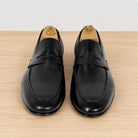 A pair of black leather penny loafers displayed on wooden shoe trees on a light wood surface against a white background.