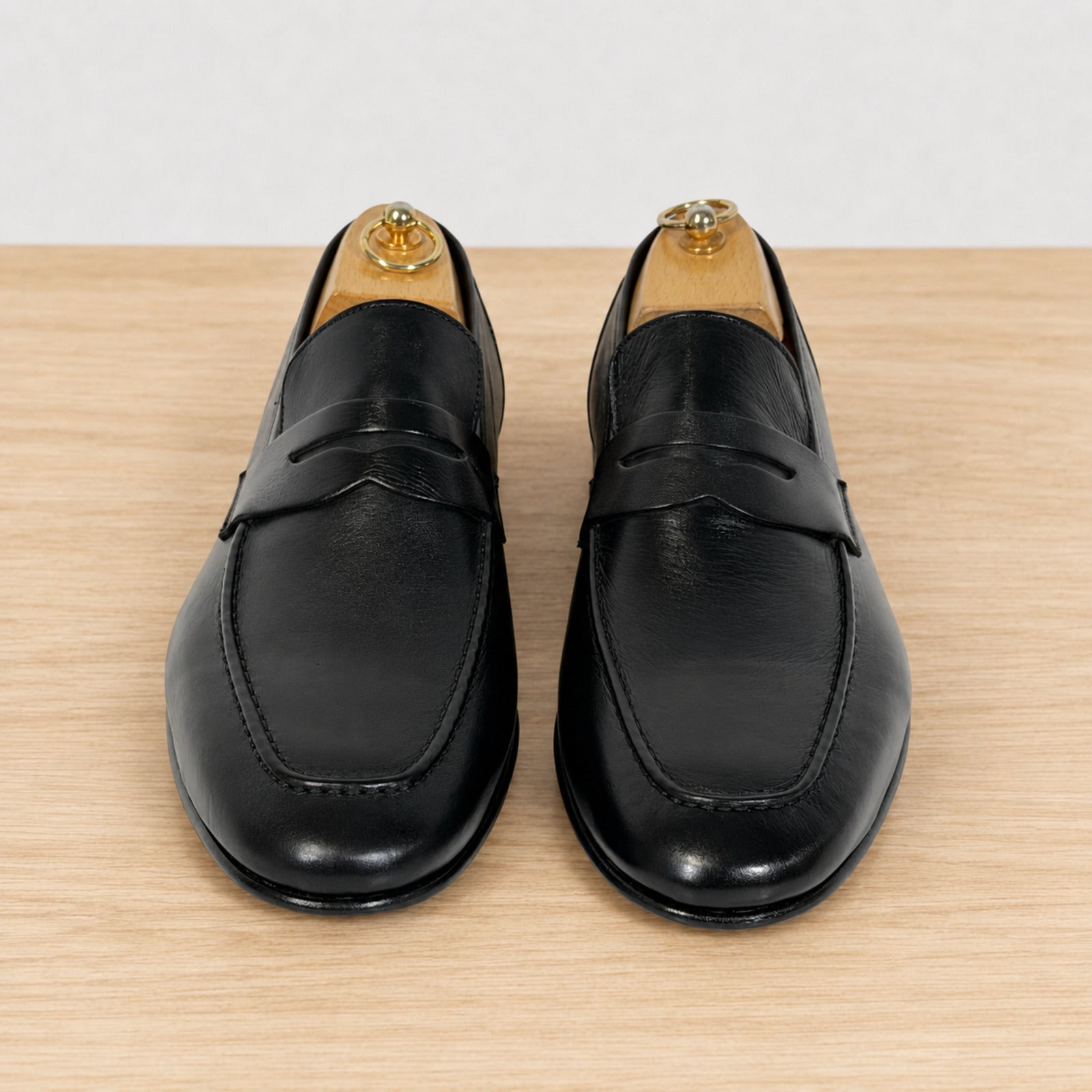 A pair of black leather penny loafers displayed on wooden shoe trees on a light wood surface against a white background.