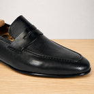 A close-up of a black leather penny loafer shoe resting on a light wood surface against a neutral light gray background.
