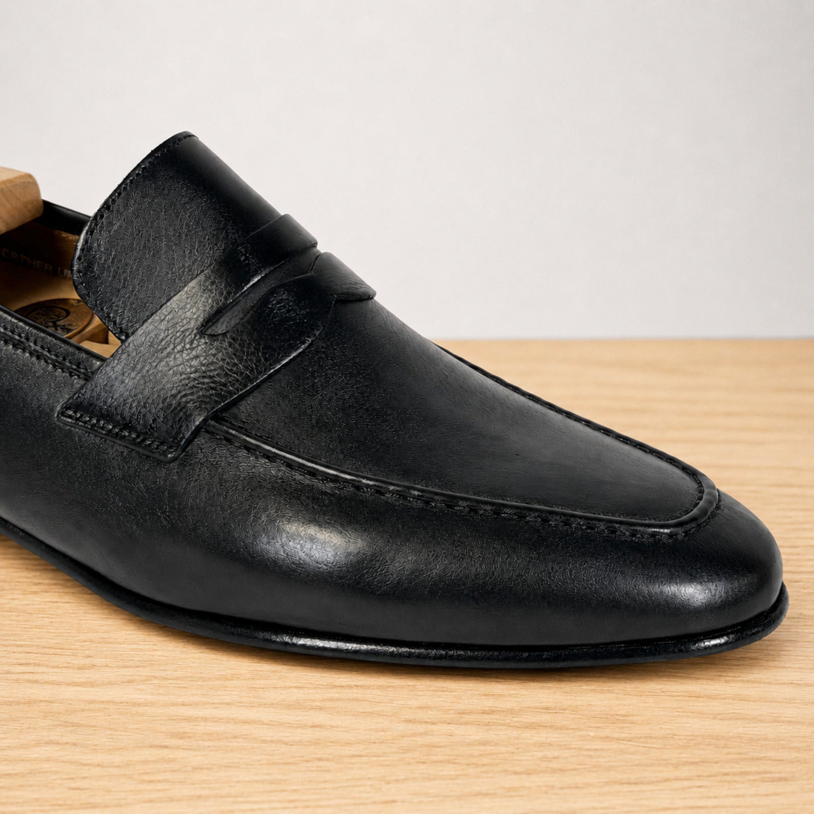 A close-up of a black leather penny loafer shoe resting on a light wood surface against a neutral light gray background.